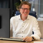 Directeur lachend achter zijn bureau met beeldscherm en toetsenbord op kantoor bij TIZ