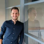 Medewerker in donker overhemd leunt lachend tegen een glazen scheidingswand op kantoor